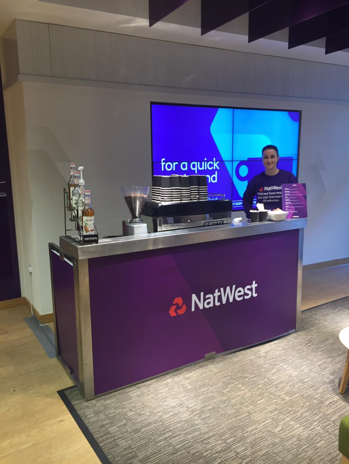 NatWest branded indoor coffee bar setup with espresso machine, syrups and a live barista station