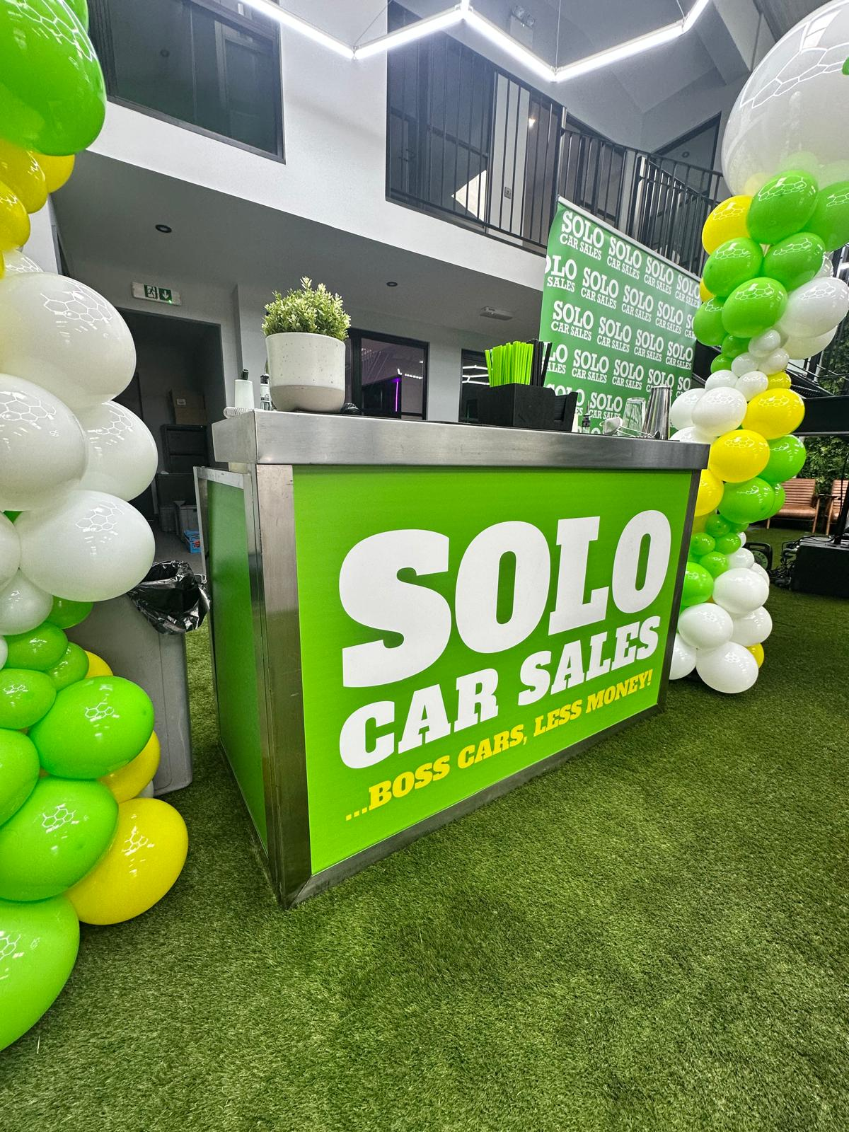 Bright green Solo Car Sales branded mobile bar installation framed by balloon displays at a launch event