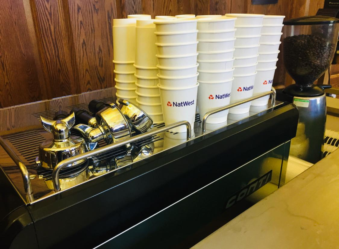 Stacks of NatWest branded takeaway cups lined up beside an espresso machine and grinder