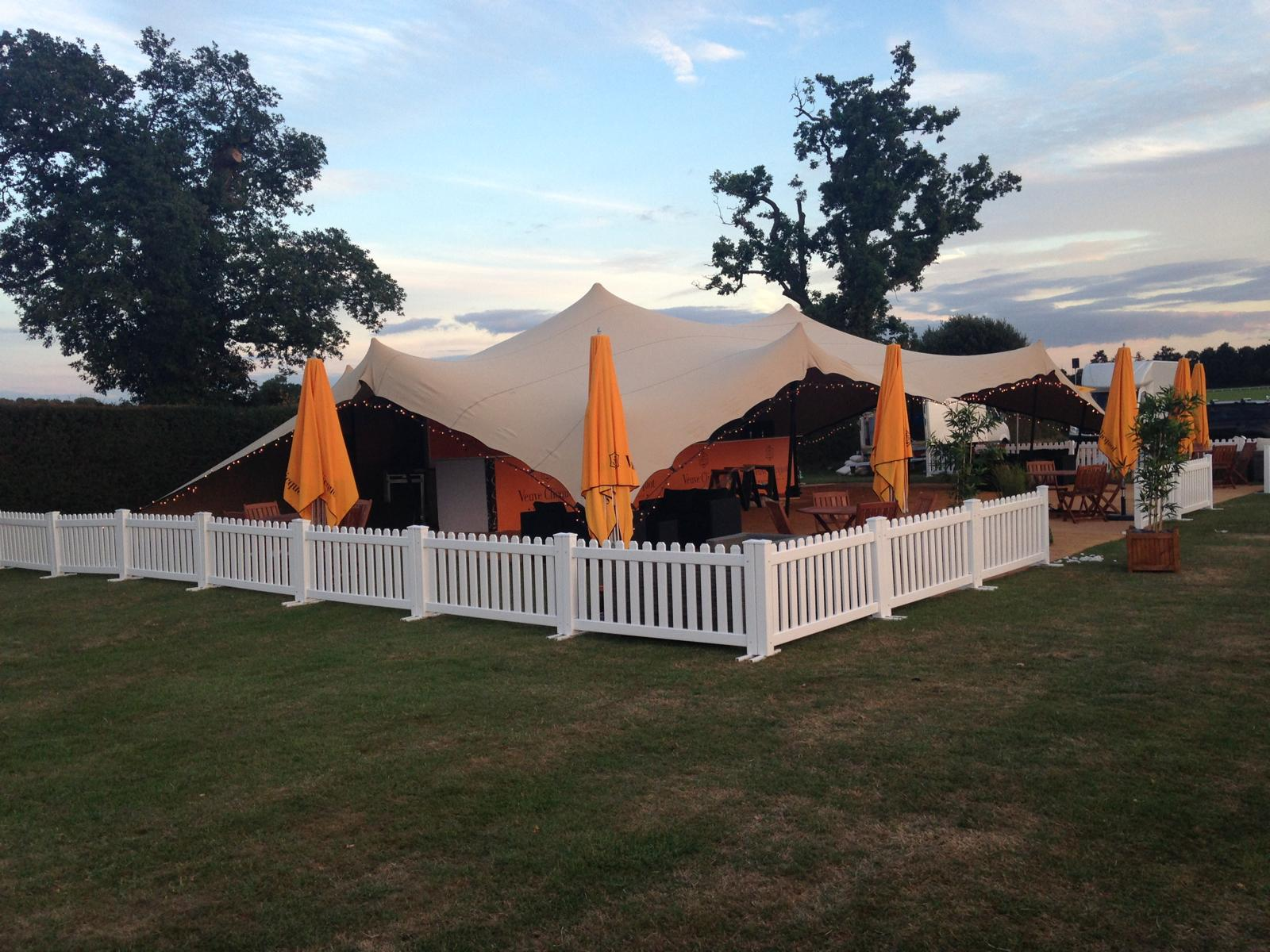Veuve Clicquot branded stretch-tent festival bar with orange parasols at dusk, set inside a white picket-fenced lawn