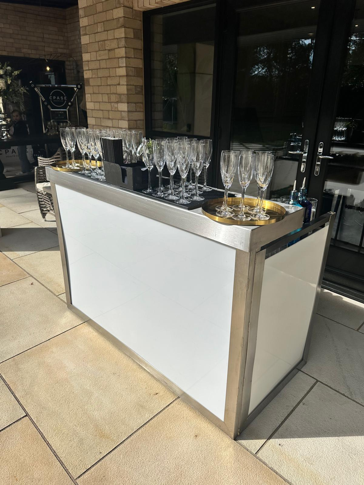 White mobile bar dressed for champagne service on an outdoor terrace with polished glassware
