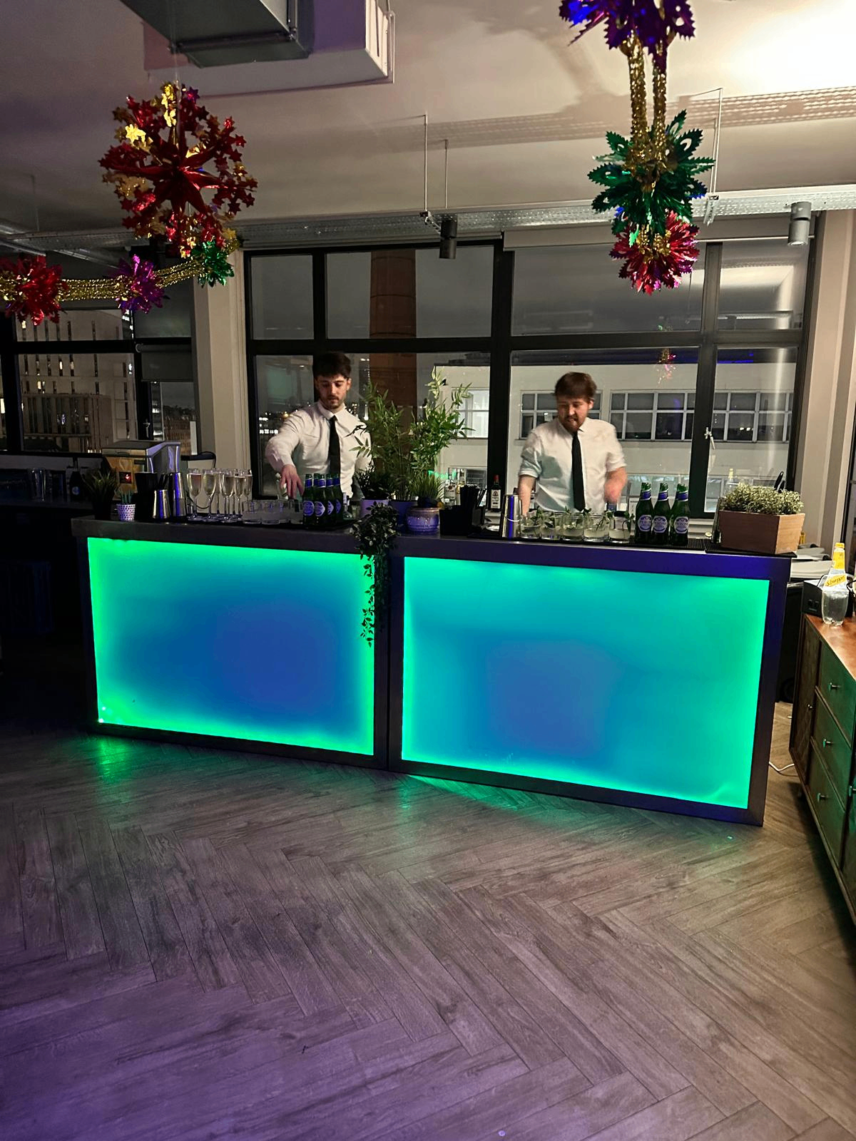 Green-lit LED mobile bar in service at a Christmas party with bartenders and festive hanging decorations
