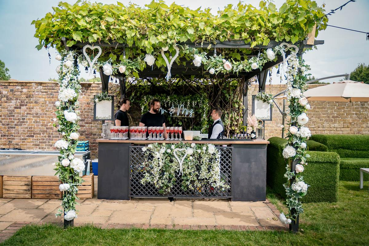 Bespoke wedding bar beneath a vine-covered pergola dressed with white florals and a Mr & Mrs neon sign in a walled garden