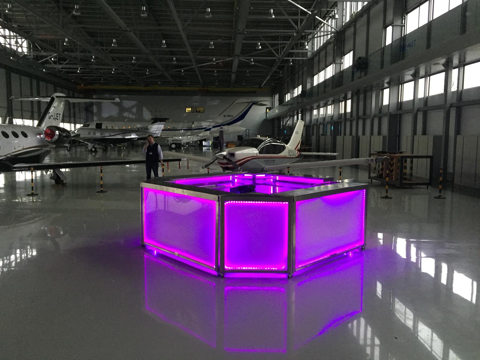 Pink-lit square island LED mobile bar set up inside a private jet hangar