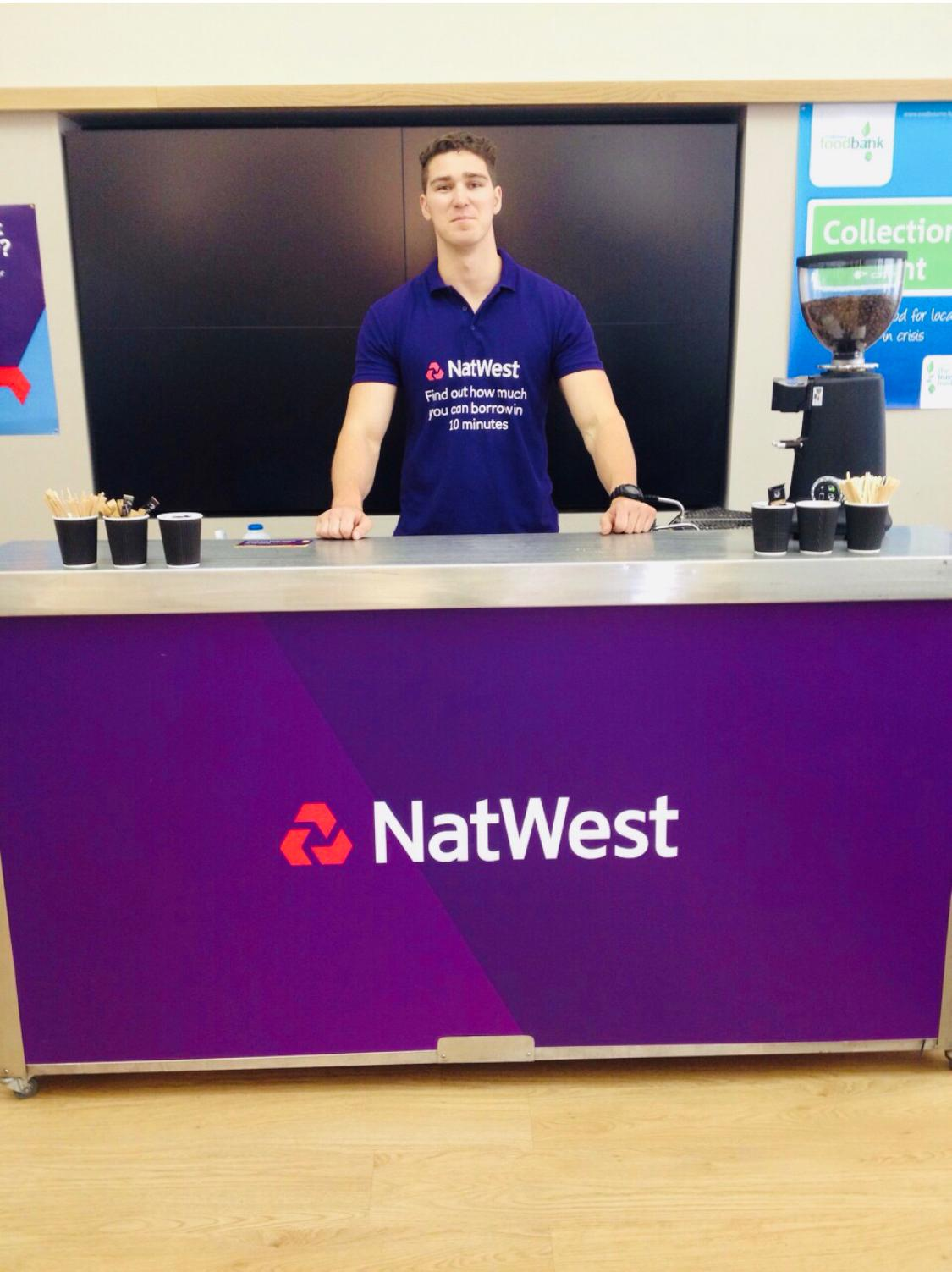 NatWest branded barista standing behind a coffee bar ready to serve fresh coffee to guests