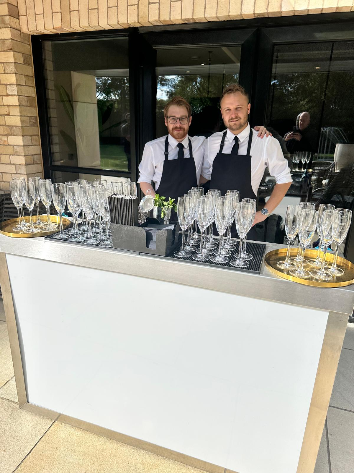 Two bartenders standing behind a white mobile bar prepared for drinks service at a private event